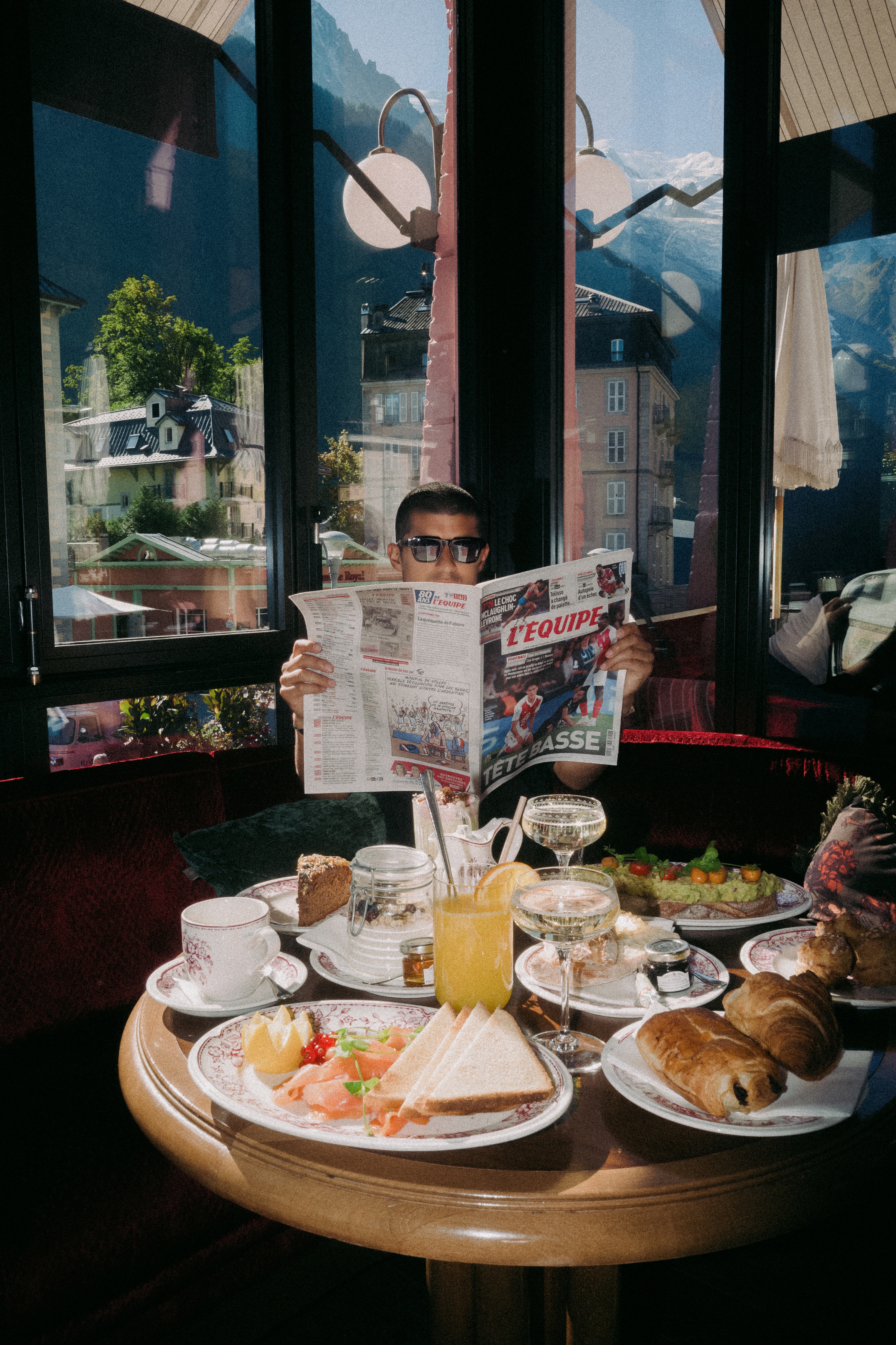 Jaime lit L’Équipe devant un petit déjeuner à la Rose du Pont, avec les montagnes derrière les fenêtres